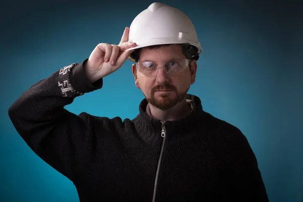Engineer with protection glasses on holding the white hardhat helmet. Studio shot of professional with work cap on.