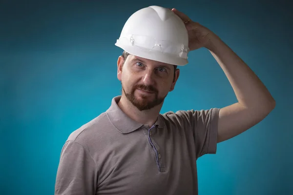 Foreman with beard holding a white safety helmet and transparent protective glasses looking playful against a studio background and blue spotlight behind