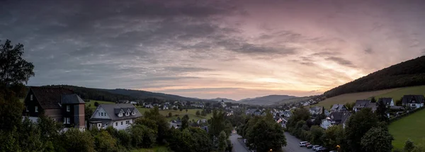 Almanya 'nın Sauerland kentindeki kış sporları bölgesinde bulunan Grafschaft adlı dağlık spa köyünün günbatımında mor ve turuncu renkli bir gökyüzü ile geniş panoramik manzarası vardır.