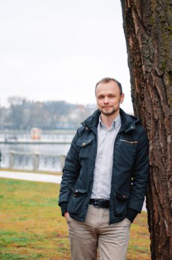 Mavi paltolu, bej pantolonlu yakışıklı bir işadamının fotoğrafı gülümsüyor ve ağacın yanında duruyor.