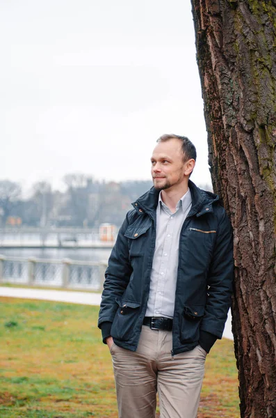 Mavi paltolu, bej pantolonlu yakışıklı bir işadamının fotoğrafı gülümsüyor ve ağacın yanında duruyor.