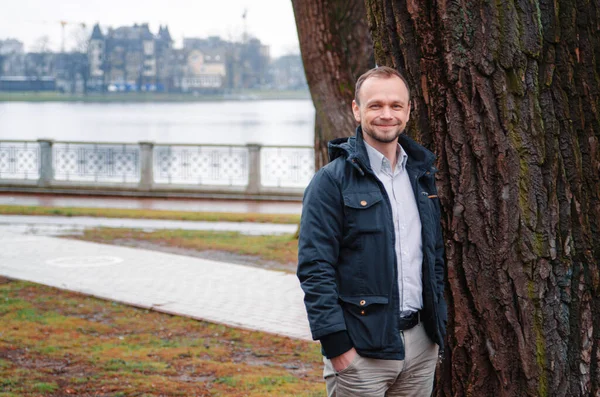 Mavi paltolu, bej pantolonlu yakışıklı bir işadamının fotoğrafı gülümsüyor ve ağacın yanında duruyor.