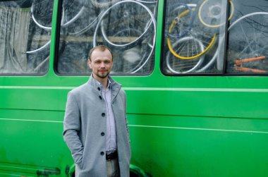 Gri paltolu ve biege pantolonlu yakışıklı bir işadamının fotoğrafı. Gülümsüyor ve yeşil otobüsün yanında duruyor.