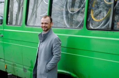 Gri paltolu ve biege pantolonlu yakışıklı bir işadamının fotoğrafı. Gülümsüyor ve yeşil otobüsün yanında duruyor.