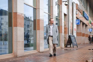 Gri paltolu ve biege pantolonlu yakışıklı işadamı gülümsüyor ve kaldırımda yürüyor.