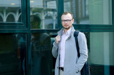 Gri paltolu ve biege pantolonlu yakışıklı işadamı gülümsüyor ve kaldırımda yürüyor.