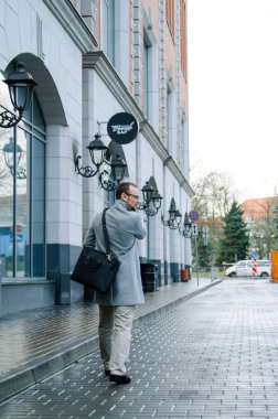Gri paltolu ve biege pantolonlu yakışıklı işadamı gülümsüyor ve kaldırımda yürüyor.
