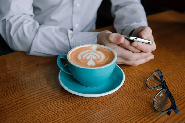 Ręce biznesmena wolnego strzelca z filiżanką kawy na stole. Niebieski kubek kawy z pianką i zdjęciem — Zdjęcie stockowe