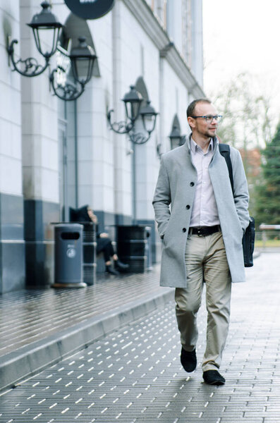 Handsome businessman freelancer in grey coat and biege trousers smiles and walking down the sidewalk