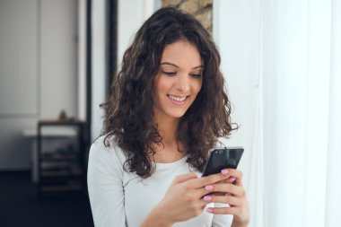 Güzel kız rahat kıyafetler içinde bir akıllı telefon kullanarak ve pencerenin yanında dururken gülümseyen