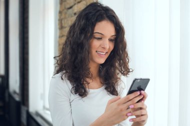 Güzel kız rahat kıyafetler içinde bir akıllı telefon kullanarak ve pencerenin yanında dururken gülümseyen