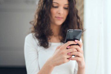 Kıvırcık kız güzel rahat kıyafetler pencerenin duran bir akıllı telefon kullanıyor