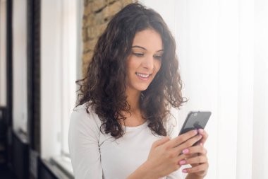 Güzel kız rahat kıyafetler içinde bir akıllı telefon kullanarak ve pencerenin yanında dururken gülümseyen