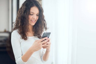 Güzel kız rahat kıyafetler içinde bir akıllı telefon kullanarak ve pencerenin yanında dururken gülümseyen