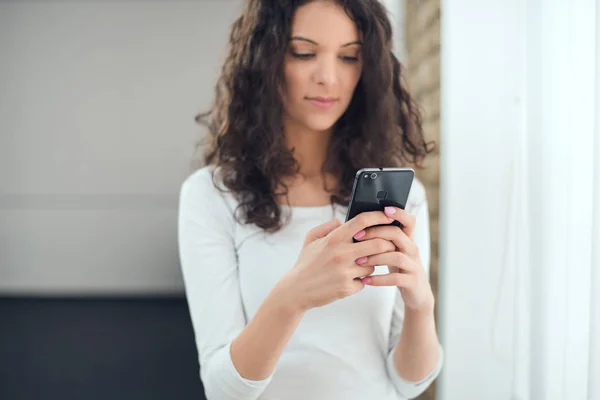 Beautiful curly woman in casual clothes is using a smart phone while standing near the window