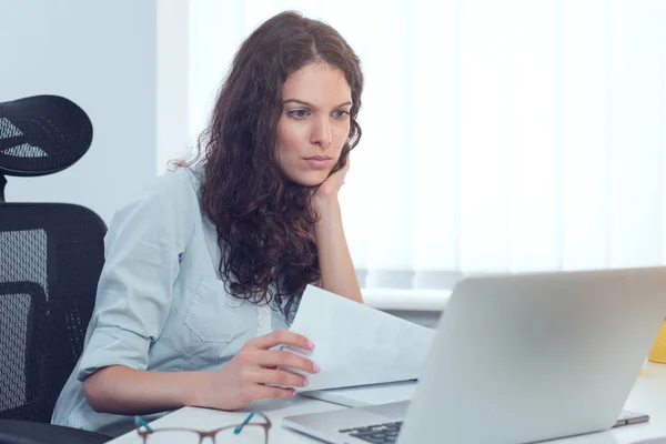 Successful serious business woman in formal wear sitting at white desk in modern office and reading on laptop, copy space