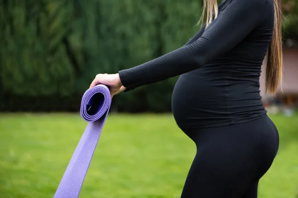 Für die Babygesundheit. junge schwangere Sportlerin steht draußen und hält den Turnteppich in der Hand und bereitet sich auf die Übungen vor. — Stockfoto