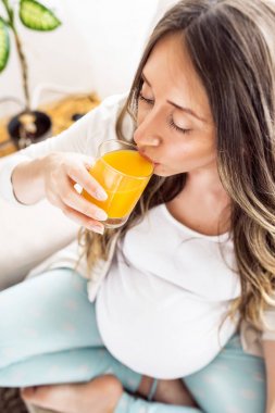 Mutlu gülümseyen zarif hamile kadın oturma odasında pencere ile evde meyve suyu smoothie içme.