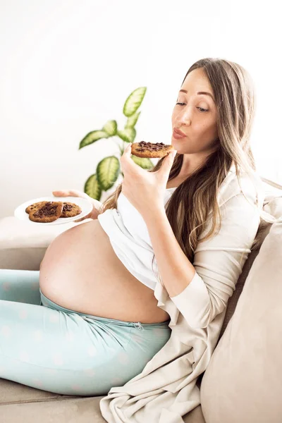 Vue latérale de la femme enceinte dégustant des cookies pour le petit déjeuner, assis près de la fenêtre regardant la caméra . Photo De Stock