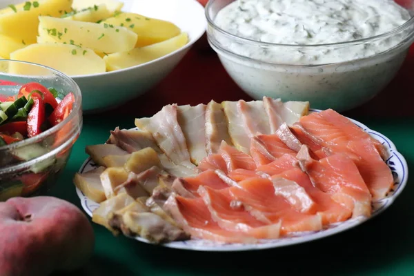 On a beautiful plate lies two types of smoked fish, sturgeon and salmon. And on the table is boiled potatoes, sauce with herbs and vegetable salad.