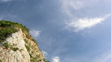 Gündüz vakti kayalık dağların uçurumlarında zaman su gibi akıyor. Arka planda mavi gökyüzü ve bulutlarla.