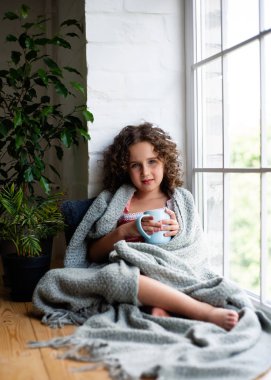 Küçük bir kız bir battaniye kaplı bir fincan çay ile pencerenin yanında oturuyor.