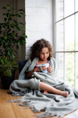 Küçük bir kız bir battaniye kaplı bir fincan çay ile pencerenin yanında oturuyor.
