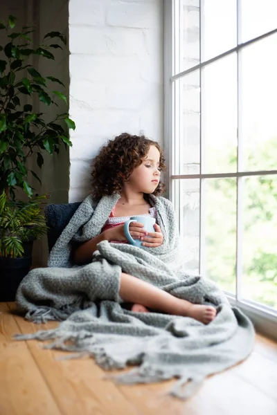 A little girl is sitting by the window with a cup of tea covered in a blanket.