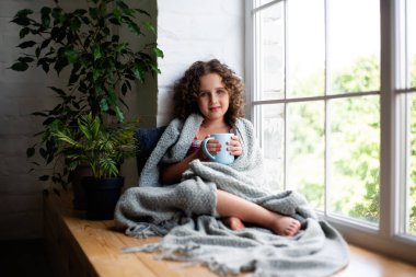 Küçük bir kız bir battaniye kaplı bir fincan çay ile pencerenin yanında oturuyor.