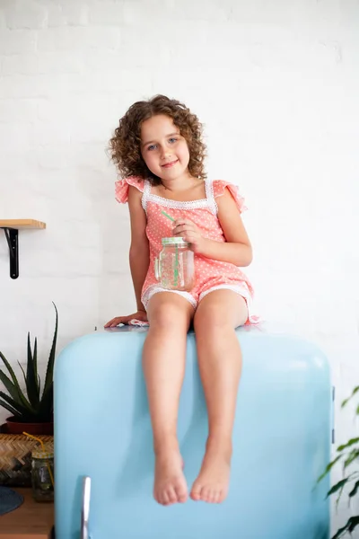 Little girl is sitting on the fridge. Sits on a beautiful retro refrigerator of blue color, stylish interior.