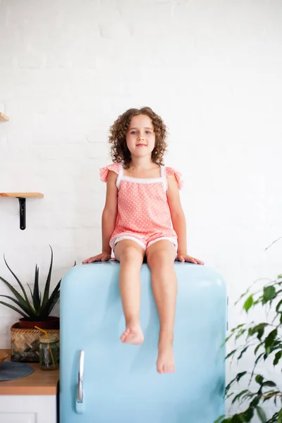 Little girl is sitting on the fridge. Sits on a beautiful retro refrigerator of blue color, stylish interior.
