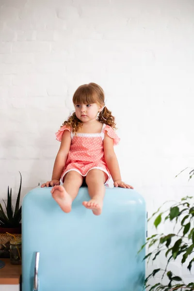 Little girl is sitting on the fridge. Sits on a beautiful retro refrigerator of blue color, stylish interior.