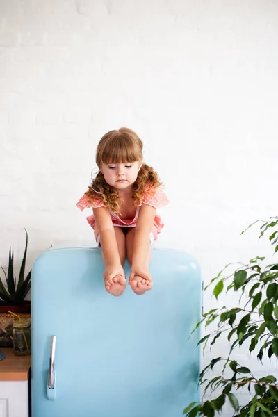 Little girl is sitting on the fridge. Sits on a beautiful retro refrigerator of blue color, stylish interior.