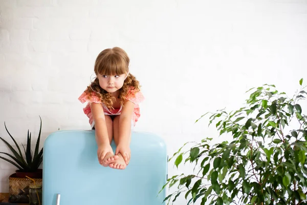 Little girl is sitting on the fridge. Sits on a beautiful retro refrigerator of blue color, stylish interior.