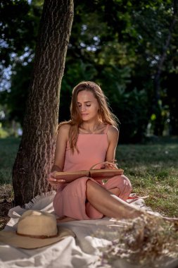 Ağacın yanında çimenlerin üzerinde dinlenen güzel kız. Kurutulmuş çiçek ve akıllı telefon bir kitap ile bir şapka içinde.