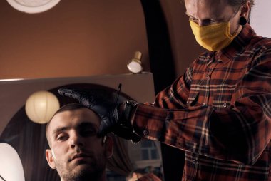 Koruyucu maske ve eldiven takmış sıradan kıyafetler giymiş bir erkek kuaför. Maskesiz müşteri. Karantinadan sonra kuaförlük. Sağlık önlemleri. Küçük bir iş. Coronavirüs salgını sonrası.