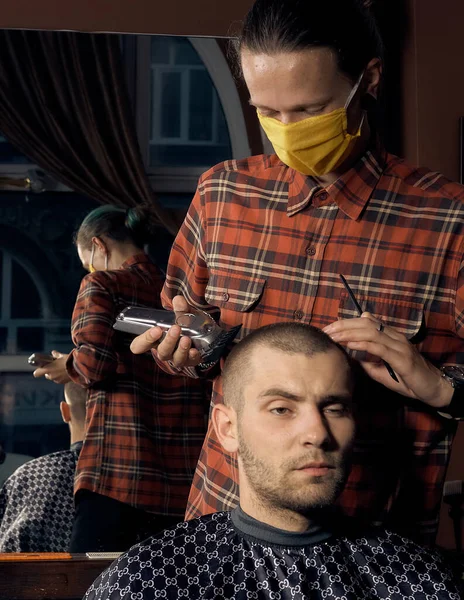 Male hairdresser in a protective mask and gloves, dressed in casual clothes. Client without a mask. Hairdressing after quarantine. Health precautions. Small business. Coronavirus pandemic aftermath.