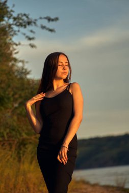 Yeşil sahil yakınında doğada güzel bir kız. Batan güneşin tadını çıkarıyor. Açık hava eğlencesi konsepti, parkta yürüyüş. Şehrin telaşından dolayı gevşeme.