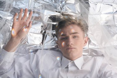 Yakışıklı bir manken gümüş folyoda tül kumaşın altında yatıyor..