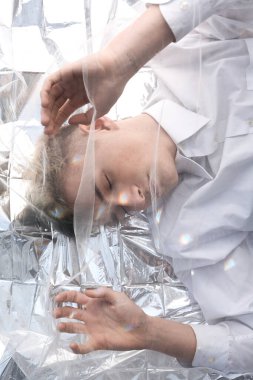 Yakışıklı bir manken gümüş folyoda tül kumaşın altında yatıyor..