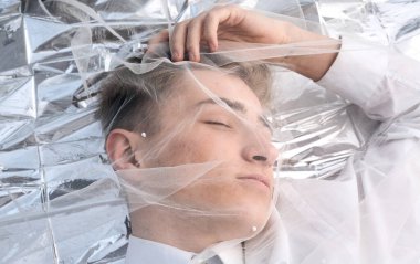 Yakışıklı bir manken gümüş folyoda tül kumaşın altında yatıyor..