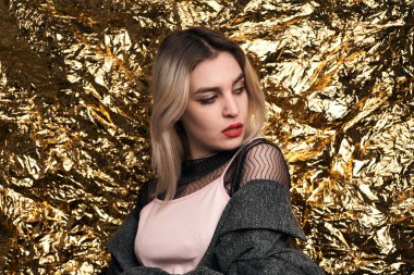 Altın bir arka planda ceketle poz veren şık bir kız..