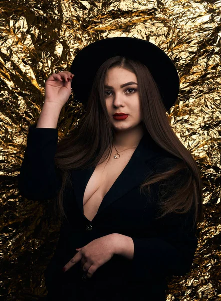 Stylish chubby girl posing in a black jacket and hat on a gold background.