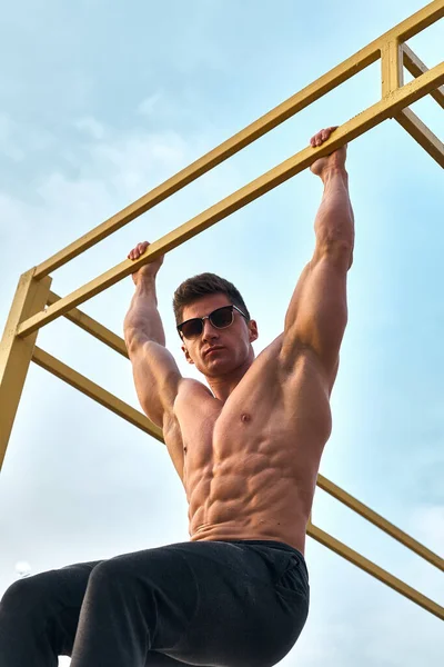 Muscular guy is training on the horizontal bar on the beach on a sunny day.