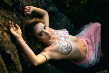 Yapışkan yapay elmaslar ve parıltılardan vücudun ve yüzün sanat yapısı. Peri perisi doğada, taşların ve suyun yanında yaşar. Fotoğrafın yumuşatma etkisi var, bir fikir olarak yapılmış..