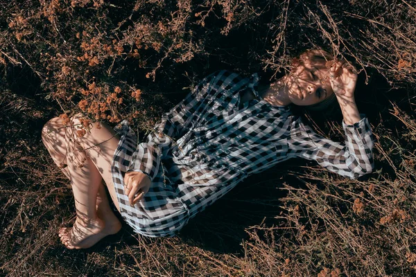 魅力的な女の子は黄色の乾燥した草の中にドレス裸足で休んでいる プレイドレスのモデル — ストック写真