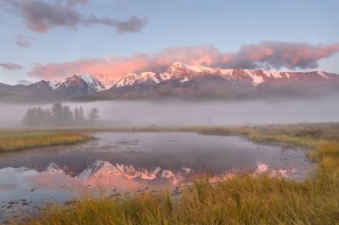 Göl, kar, yeşil ağaçlar, çimenler, mavi gökyüzü, bulutlar ve yansımaları ile kaplı dağları üzerinden güzel bir sis ile şaşırtıcı pembe gündoğumu