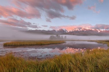 Göl, kar, yeşil ağaçlar, çimenler, mavi gökyüzü, bulutlar ve yansımaları ile kaplı dağları üzerinden güzel bir sis ile şaşırtıcı pembe gündoğumu