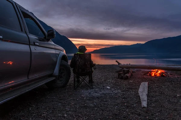 The tourist sits on the shore of the lake near a car and a fire and watches a beautiful sunset over the lake and mountains