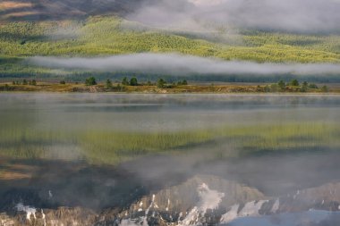 Yeşil orman, tepeler, ağaçlar ve güzel sisle kaplı dağların yansımalarıyla büyüleyici bir manzara sabahın erken saatlerinde gölün pürüzsüz sularında.